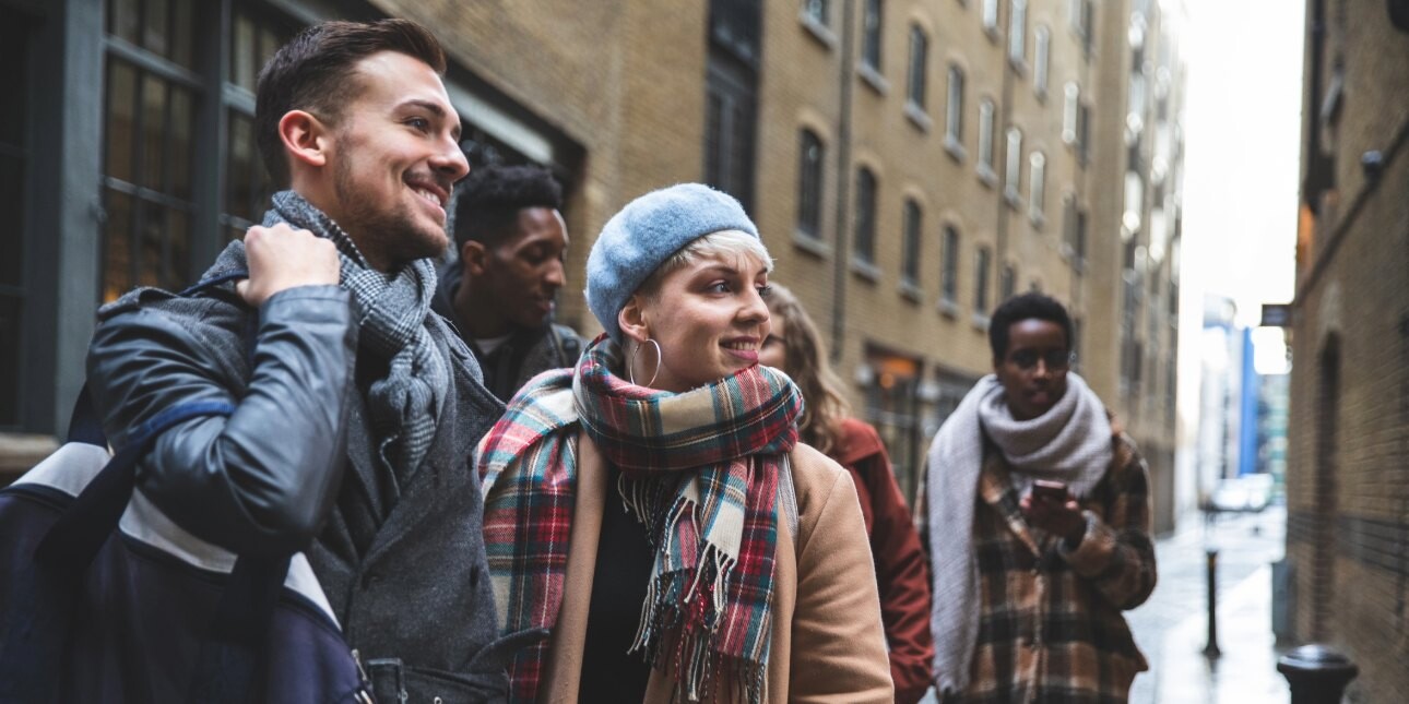 On a London street on a cold day, a group of young adults huddle while wearing scarfs and winter coats